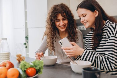 Two teen girls enjoying breakfast together at the kitchen table, happily sharing moments while looking at a smartphone and eating healthy food