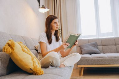 Young woman relaxing on her comfortable sofa reading a book in her living room enjoying her free time