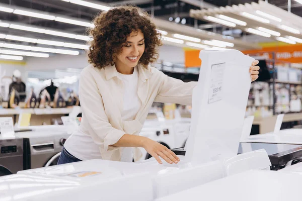 Smiling woman opening washing machine checking features in home appliance store, choosing new washer for her apartment