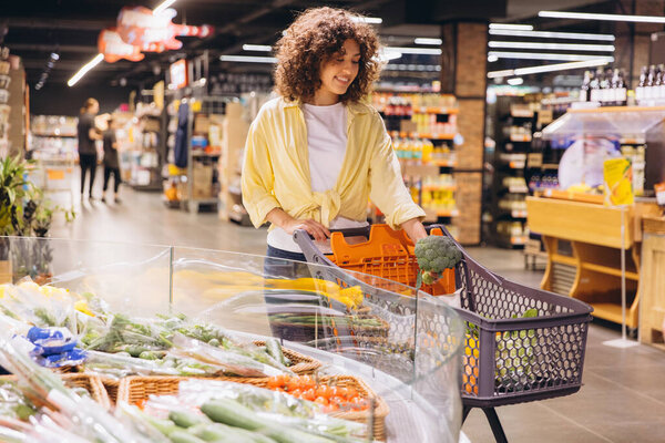 Young woman buying groceries, choosing fresh vegetables in supermarket, putting broccoli in shopping cart