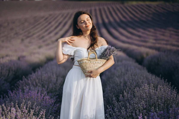 Young woman with closed eyes holding wicker basket and smelling lavender bouquet in beautiful blooming field at sunset