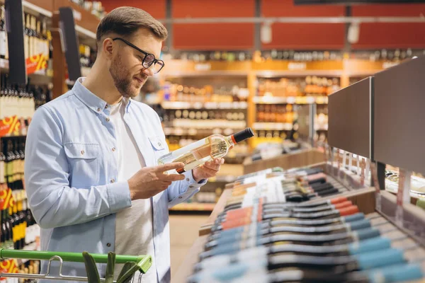 Man reading label of white wine bottle while shopping in supermarket drinks aisle