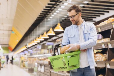 Customer wearing gloves choosing bread in supermarket bakery section with shelves full of bread in background
