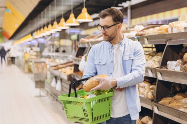 Man choosing bread from shelf in supermarket bakery section wearing gloves and holding shopping basket