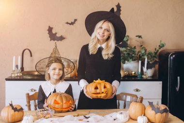 Mother and daughter wearing witch costumes holding carved pumpkins during Halloween party in decorated kitchen