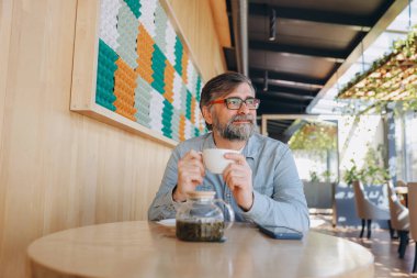 Bearded senior man sipping tea in a modern cafe, gazing thoughtfully out the window, enjoying a peaceful moment of relaxation