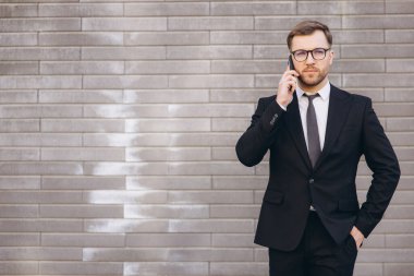 Confident businessman wearing suit and tie, using smartphone in front of gray brick wall