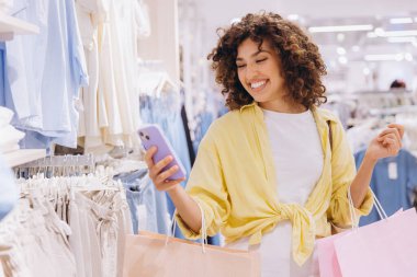Happy customer using mobile phone and carrying shopping bags while buying clothes in fashion store