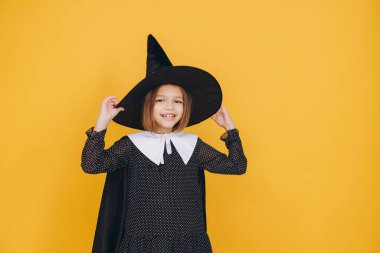 Studio portrait of a cheerful girl wearing witch costume holding her hat posing on a yellow background for Halloween