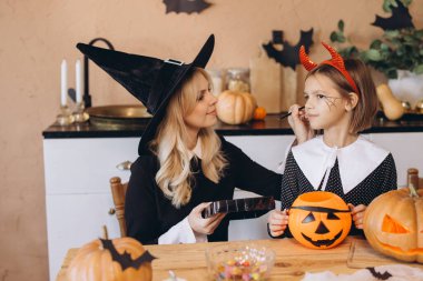 Mother wearing witch costume applying Halloween makeup on her daughter's face, sitting at a table with pumpkins and sweets