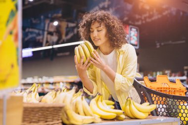 Customer enjoying the aroma of ripe bananas while selecting fresh produce in a vibrant supermarket, surrounded by colorful grocery options