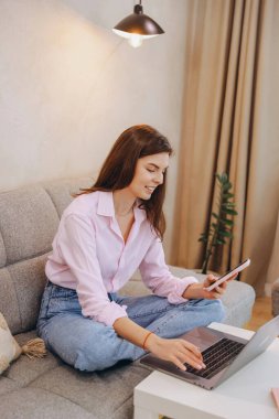 Freelancer managing her business using laptop and mobile phone, comfortably sitting on the sofa in her living room