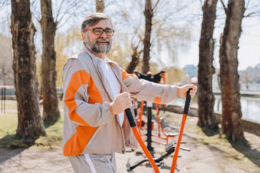 Happy bearded senior man wearing sportswear exercising using equipment at an outdoor gym in a park near a lake