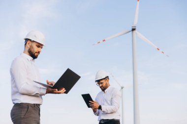 Two engineers working with laptop and tablet near wind turbines, checking data for sustainable energy production