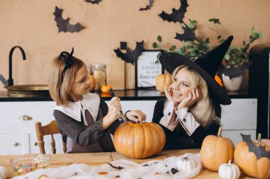 Mother wearing a witch hat and daughter happily carving a pumpkin together for their festive Halloween celebration at home