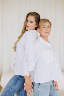 Mother and daughter wearing white shirts and jeans standing back to back holding hands, looking over their shoulders