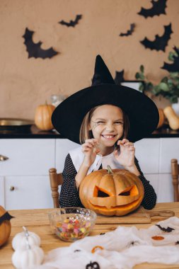 Girl wearing witch costume preparing jack o' lantern with candies on wooden table in kitchen decorated for Halloween