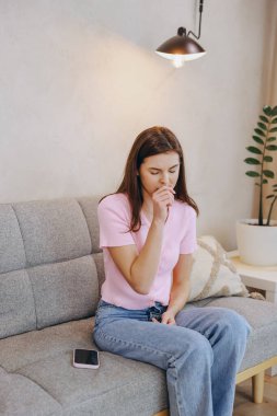 Young woman feeling unwell, coughing and covering her mouth with hand while sitting on sofa in living room