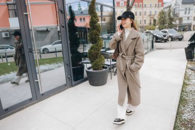 Young manager wearing a beige coat and black cap, enjoys a phone call while walking near modern buildings in a city setting