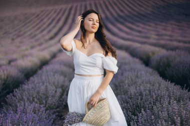 Beautiful model touching her hair with a wicker bag in her hand, enjoying the scent of lavender in a large field on a sunny summer day