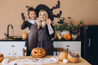 Mother and daughter making funny faces and wearing witch costumes are preparing carved pumpkins and candies for Halloween party