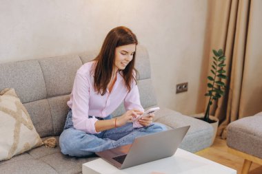 Smiling woman sitting comfortably on a sofa, using a laptop and smartphone to work remotely from home while enjoying a relaxed atmosphere