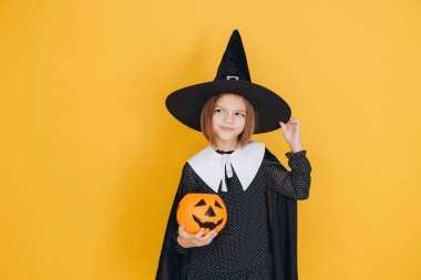 Little girl wearing Halloween witch costume holding a pumpkin bucket and slightly adjusting her hat on a yellow background