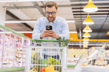 Customer using smartphone and smiling while shopping for groceries in a supermarket with a full cart