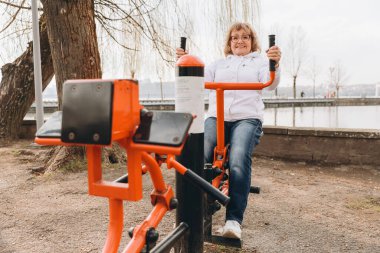 Happy senior woman exercising on outdoor gym equipment by the lake, enjoying a healthy and active lifestyle