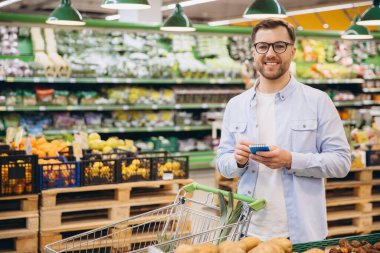 Smiling man writing grocery list while shopping in supermarket with shopping cart