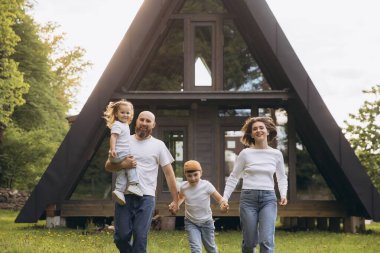 Cheerful family with two children running toward camera in front of their modern a frame house in the countryside