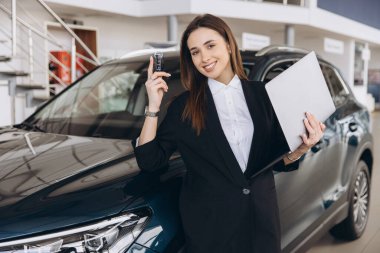 Saleswoman is showing car key and holding laptop while standing next to a new car in a dealership