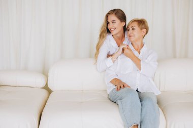 Happy mother and daughter holding hands and smiling, enjoying a peaceful moment together on a comfortable white sofa in a brightly lit room