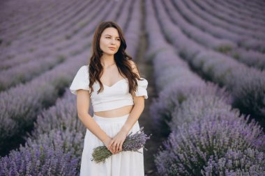 Young woman wearing a flowing white dress, holding a fragrant lavender bouquet while surrounded by a stunning lavender field in bloom