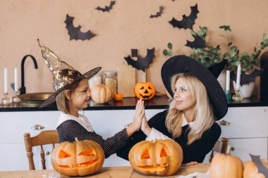 Mom and daughter with witch hats celebrating Halloween by giving high five in their kitchen over carved pumpkins
