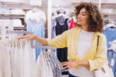 Smiling woman with curly hair selecting stylish clothes from hangers in a vibrant fashion store, enjoying the shopping experience