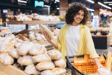 Smiling curly haired woman choosing bread from shelf in supermarket bakery section, holding shopping cart