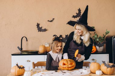 Mother wearing witch hat and daughter carving pumpkins for Halloween party, kitchen decorated with bats