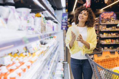 Young woman choosing a bottle of milk in supermarket refrigerator shelf pushing a shopping cart