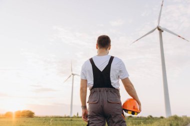Engineer holding his helmet and admiring wind turbines in a field at sunset, representing sustainable energy and ecological responsibility