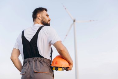 Engineer holding an orange helmet, gazing at a wind turbine against a backdrop of clear blue sky, emphasizing sustainable energy solutions