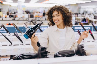 Smiling woman with curly hair selecting a hairdryer while browsing through the electronics store, enjoying the shopping experience