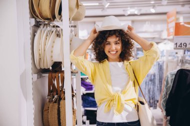 Smiling young woman trying on a stylish white hat while shopping in a trendy fashion store, enjoying the experience of selecting new accessories