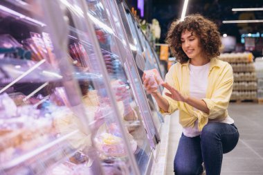 Smiling curly haired woman crouching and choosing packaged food from refrigerated section in supermarket