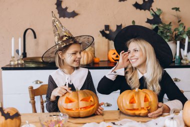 Mother and daughter, dressed in witch costumes, joyfully carving pumpkins together for a festive Halloween celebration at home