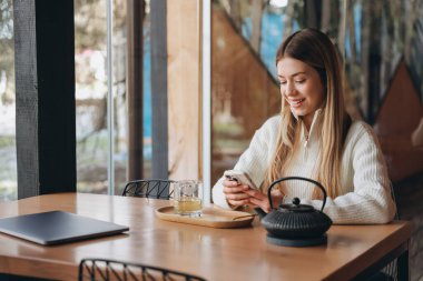 Freelancer working remotely from cafe, enjoying tea and using smartphone, with laptop on wooden table