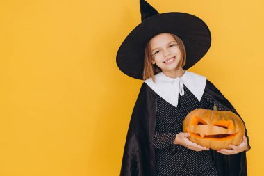 Portrait of a smiling little girl wearing witch costume holding carved pumpkin on yellow background celebrating Halloween