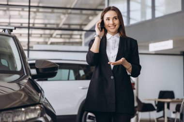 Smiling saleswoman engaging in phone conversation while presenting a new car to a customer in a modern car dealership showroom