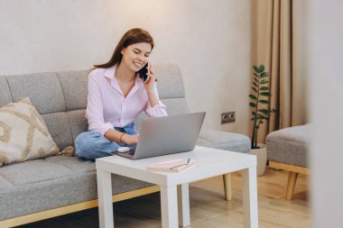 Freelancer working remotely from home, using laptop and mobile phone while sitting comfortably on sofa