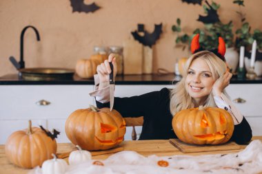 Young blonde woman wearing devil horns, carving pumpkins for a Halloween party celebration, surrounded by festive decorations in the kitchen